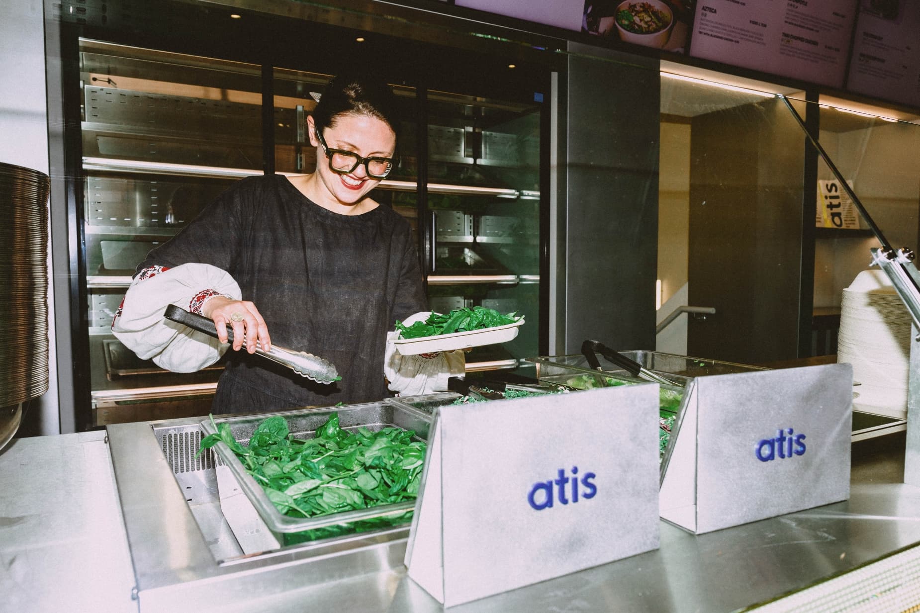 Atis staff member smiling and serving fresh greens at the counter